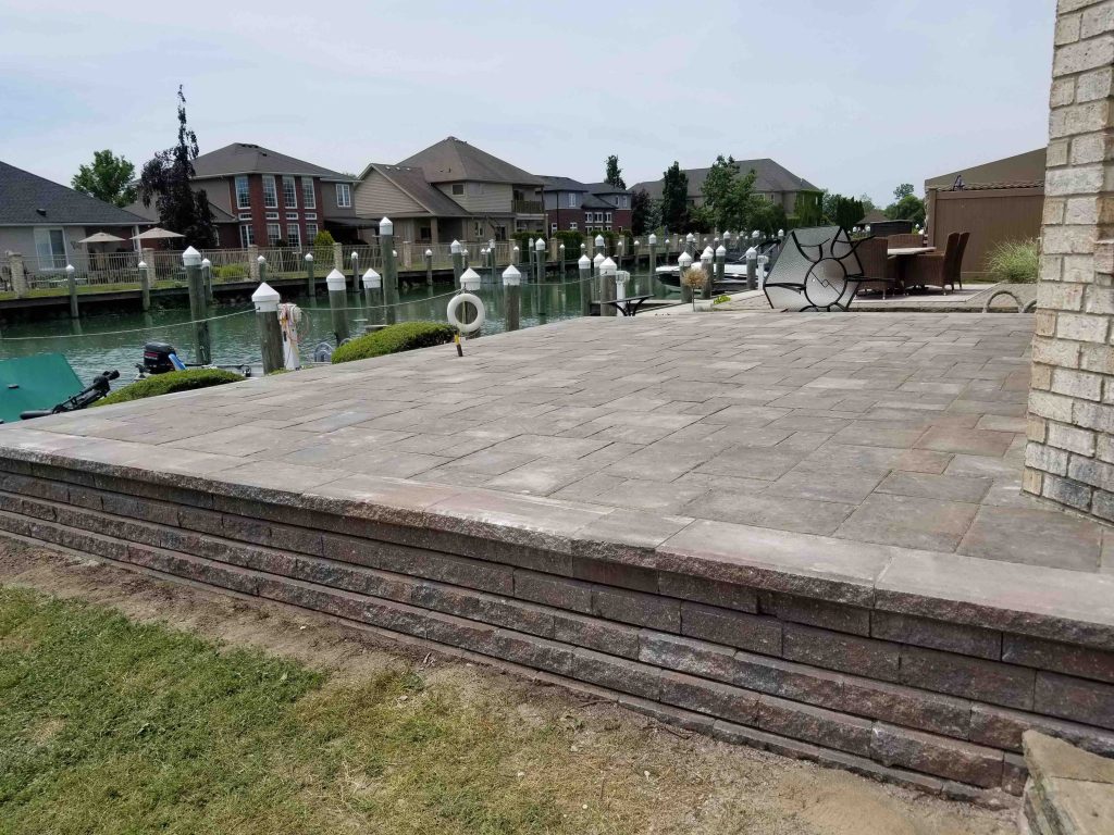 Elevated stone patio with a retaining wall beside a waterfront, featuring outdoor furniture and surrounded by a residential neighborhood.